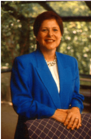 Janice Ballice is wearing a blue suit with a white shirt. She stands behind a chair.