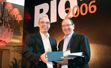 Two men in business attire stand together for a photo in front of a sign with Rio 2006 written on it.