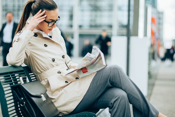 Young woman in business wear sitting on a bench in modern urban setting and reading a newspaper.