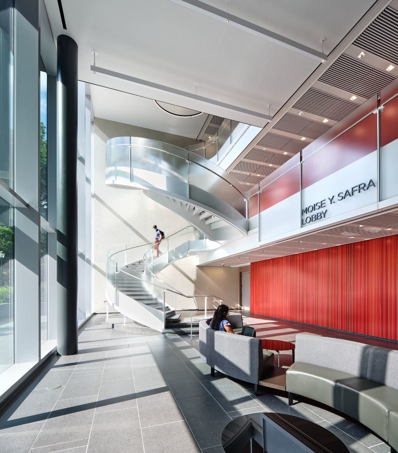 The main lobby of the Academic Research Building, with a student sitting in a modern couch and another student walking down the lobby's centerpiece spiral staircase.