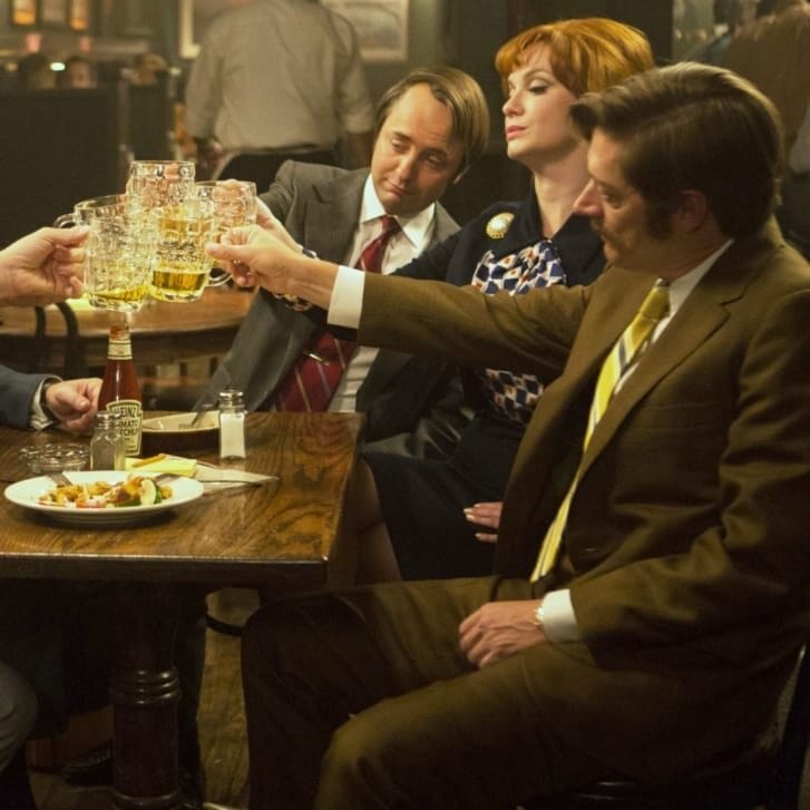 Actors in a scene from Mad Men clink glasses at a dinner table.
