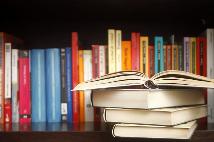 Stack of books on a wooden library shelf, the one on top open , and multicolored book spines in a row in the background.