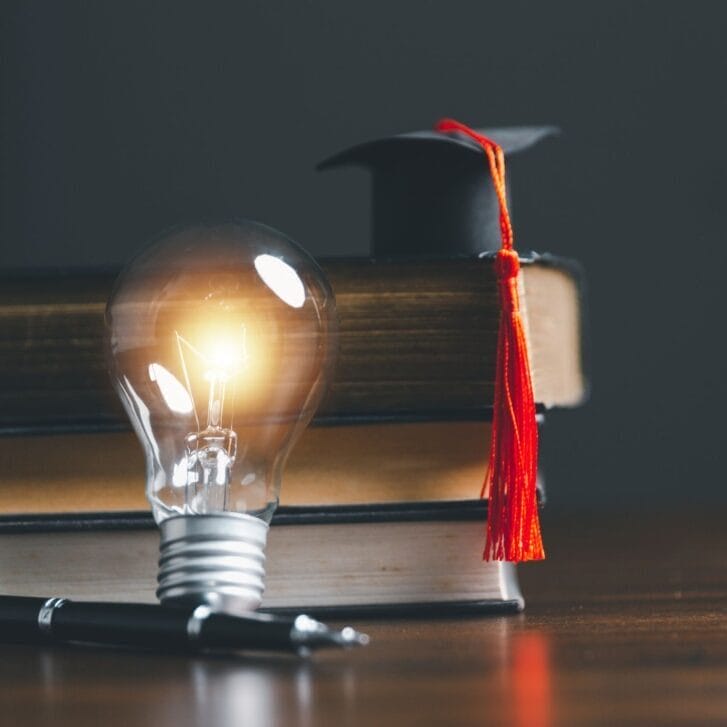 Conceptual photo of a lit lightbulb leaning against a stack of books with a graduation cap on top of the stack.
