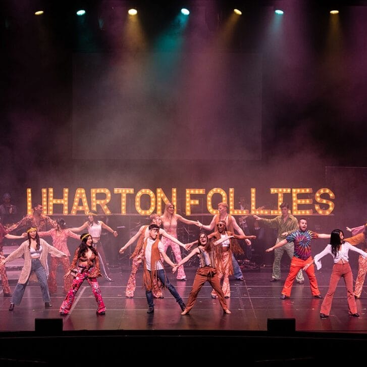 Nineteen members of the Follies dance on stage in front of a lit sign saying 