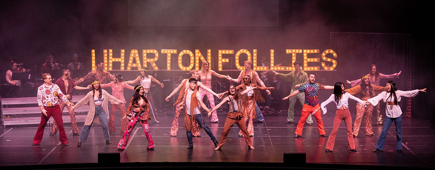 Nineteen members of the Follies dance on stage in front of a lit sign saying 