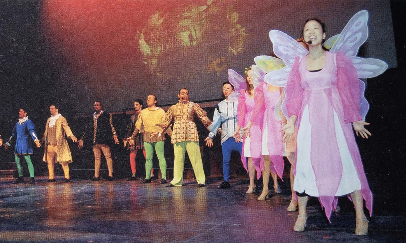 Members of the Follies perform on stage in a fantasy-like scene, with some members dressed as fairies and others in medieval garb.