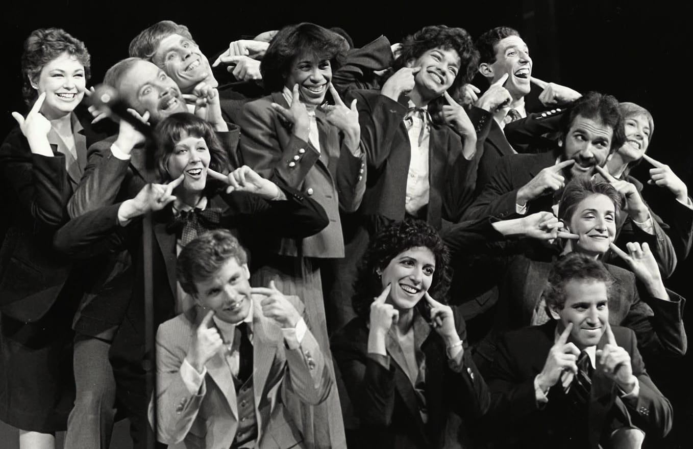 Members of the Wharton Follies wearing business suits and pointing to their own smiles in a past performance.