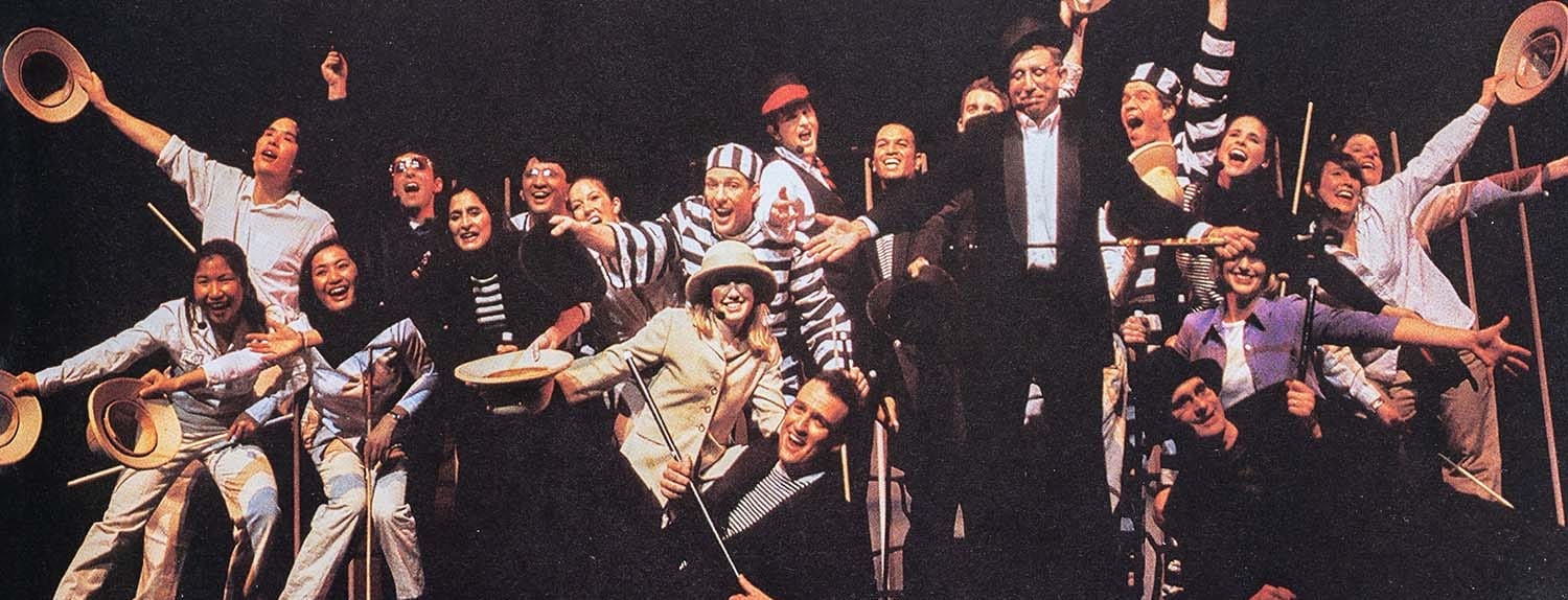Many alumni dancing on stage for a past Follies performance, some with canes and hats, others in jail suits, and some in plain business attire.