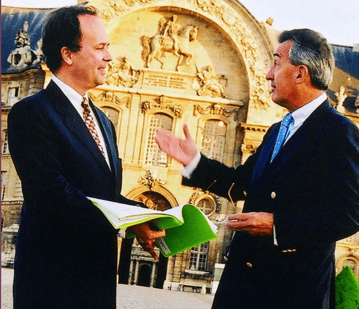 Two men in suits stand outside speaking to one another, one man holding a green notebook.