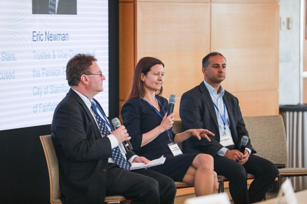 Three people in business attire sit in front of an audience holding microphones for a panel discussion.