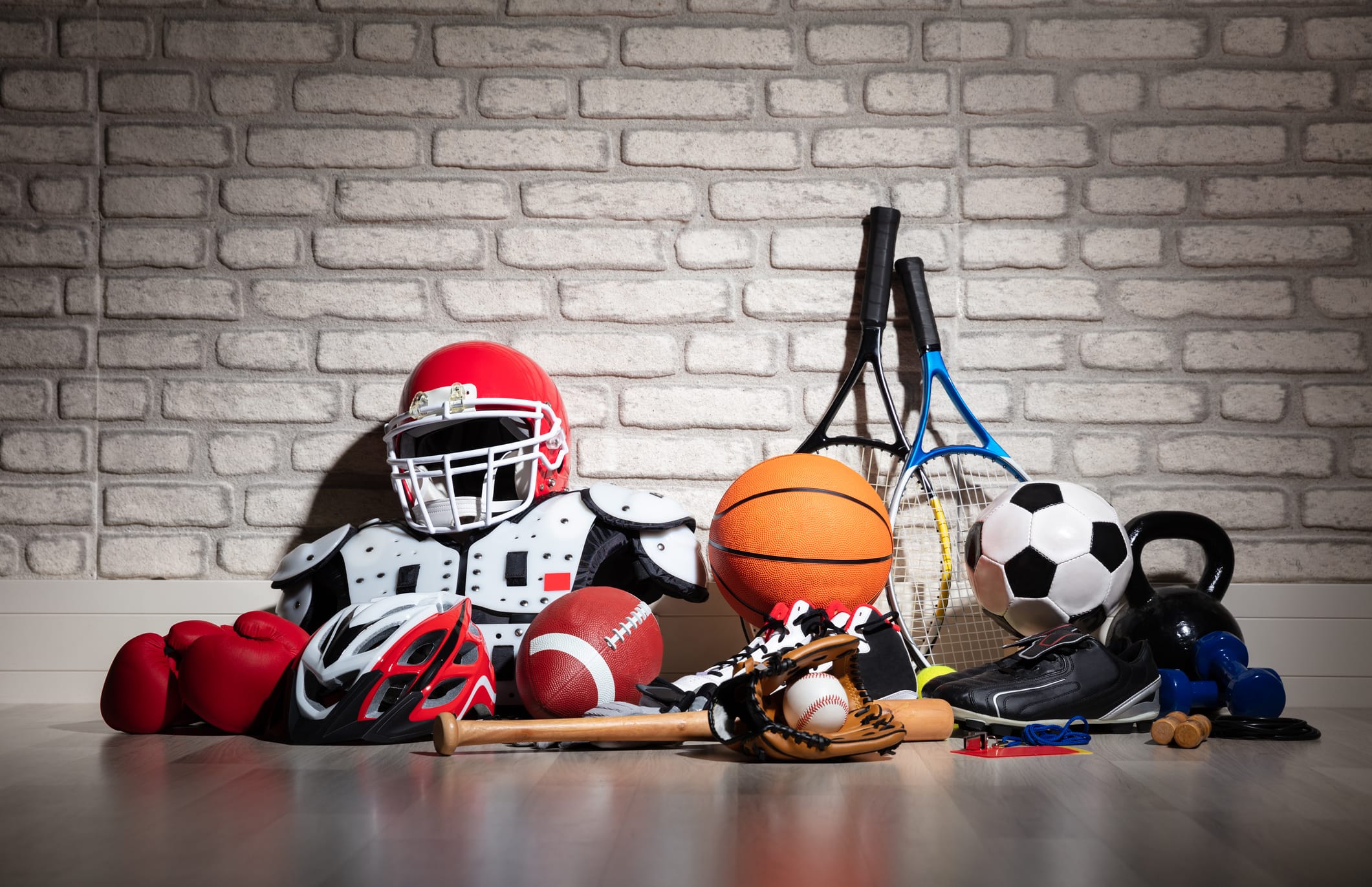 Sports equipment is laid up against wall. From left to right: boxing globes, bike helmet, football pads and helment, football, baseball bat, baseball glove and baseball, basketball, rackets, soccerball, and dumbell weights