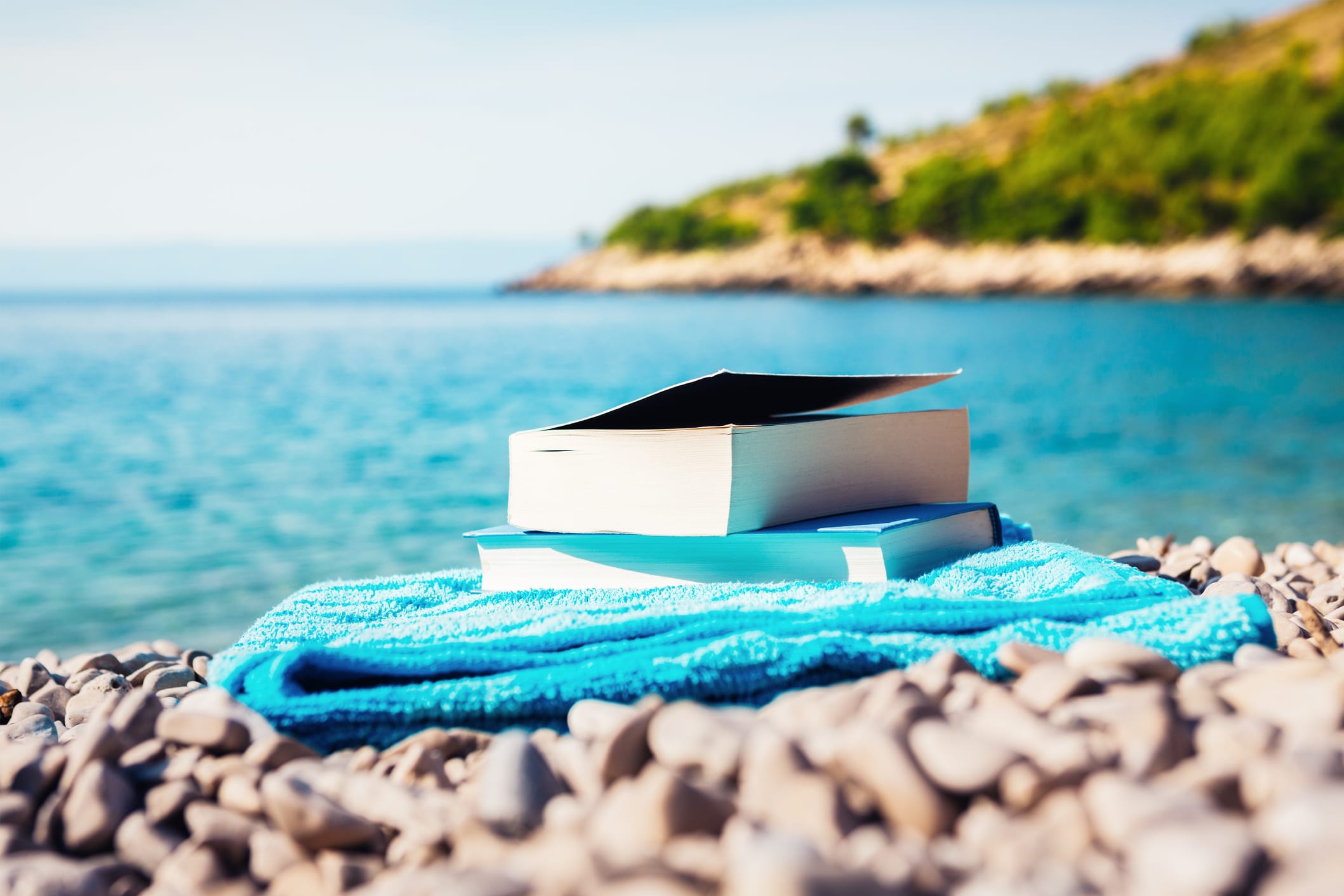 At a beach, two books sit on top of a blue towel on a rocky shore. The water is in the background, with trees blurred in thee distance.
