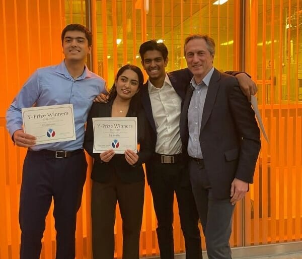 Three students and the executive director of the Mack Institute pose together smiling, with two of the students holding paper awards.