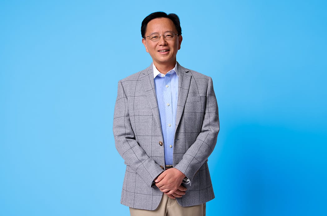 A man in a gray blazer, light blue shirt, and tan khakis poses for a photo in front of a light blue background.