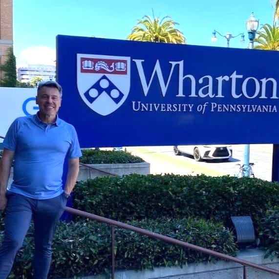 A man in a blue polo stands in front of a Wharton sign on the Wharton campus in San Francisco.