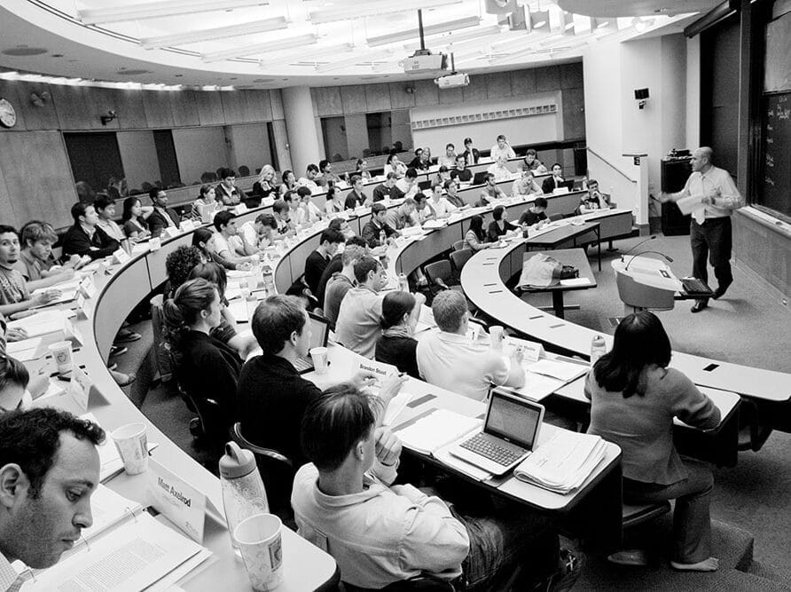 Students seated in a lecture hall with desks and a professor teaching in front of a whiteboard at the front.