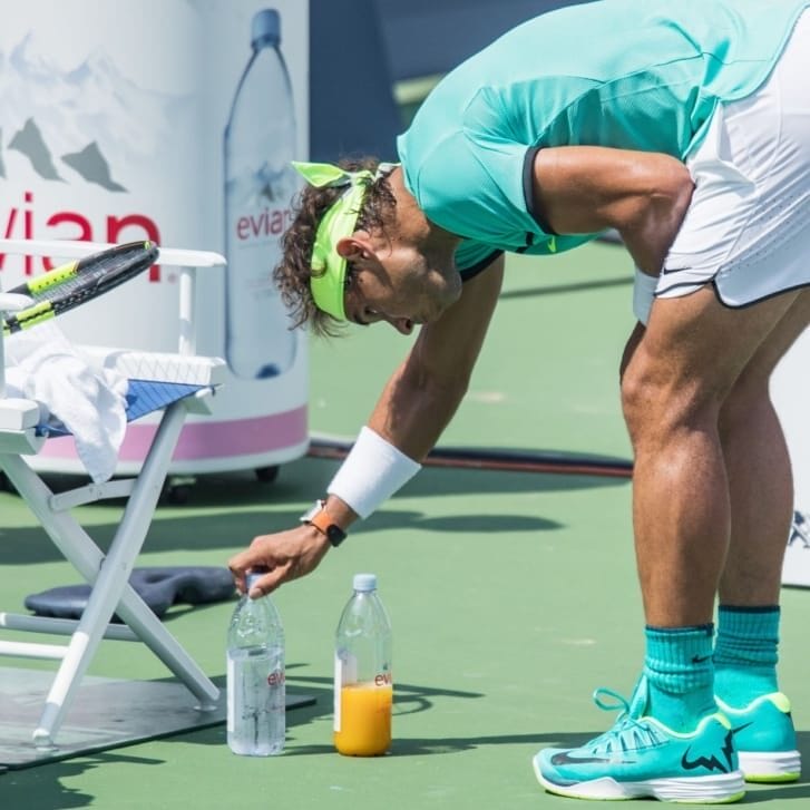 A tennis player stares intently at two bottles that he adjusting on the ground in front of a courtside chair.