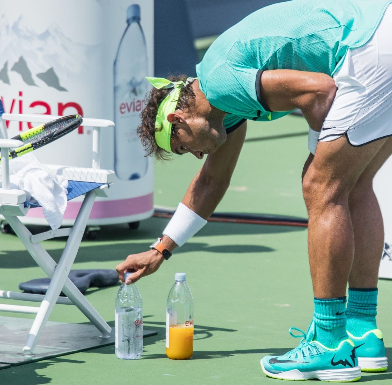A tennis player stares intently at two bottles that he adjusting on the ground in front of a courtside chair.