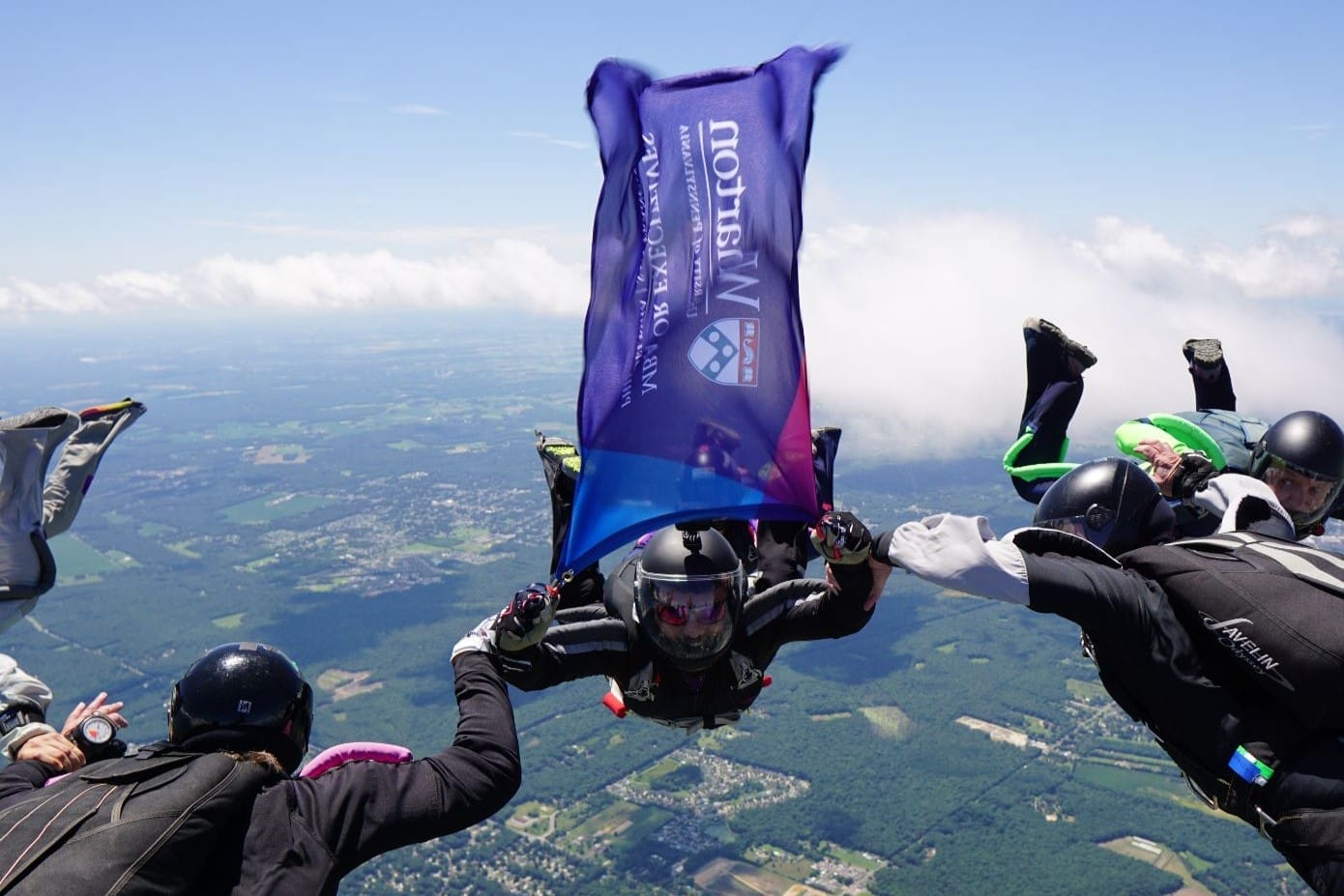 Skydivers float in formation high above the ground, with one skydiver holding up a billowing Wharton flag.