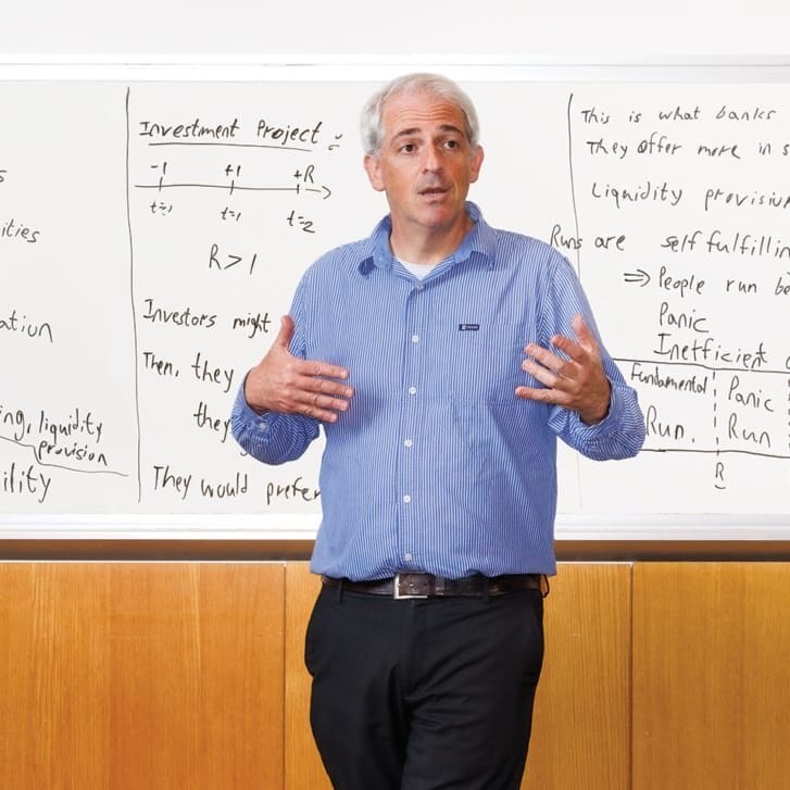 A man in a white and blue striped shirt and black pants gestures in front a whiteboard filled with notes explaining what bank runs are.