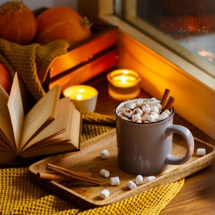 A book laying on a table amid small pumpkins, two candles, and a mug with a hot drink in it.