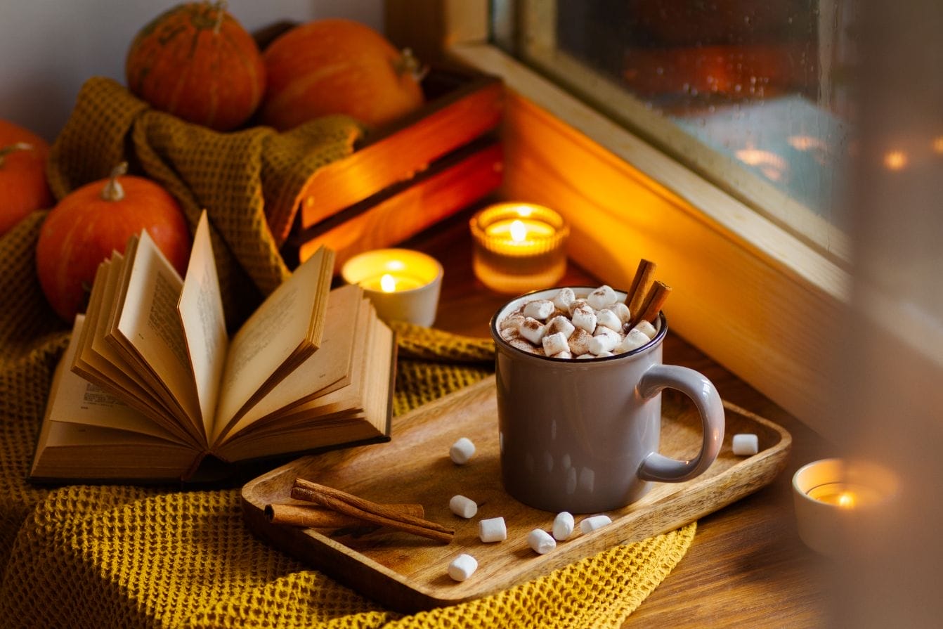 A book laying on a table amid small pumpkins, two candles, and a mug with a hot drink in it.