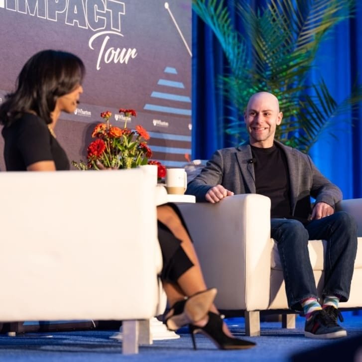 Wharton professor Adam Grant seated with Dean Erika James on stage before a blue backdrop branded for the Wharton Impact Tour.