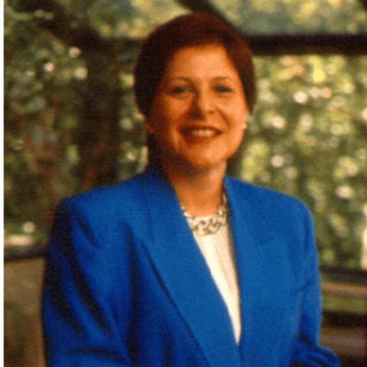 Janice Ballice is wearing a blue suit with a white shirt. She stands behind a chair.