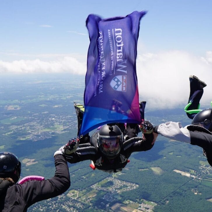 Skydivers float in formation high above the ground, with one skydiver holding up a billowing Wharton flag.