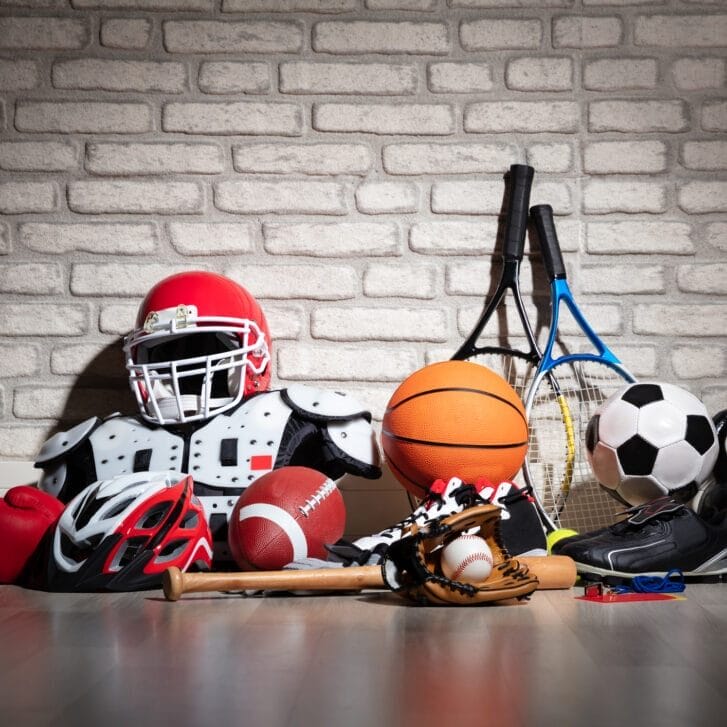 Sports equipment is laid up against wall. From left to right: boxing globes, bike helmet, football pads and helment, football, baseball bat, baseball glove and baseball, basketball, rackets, soccerball, and dumbell weights