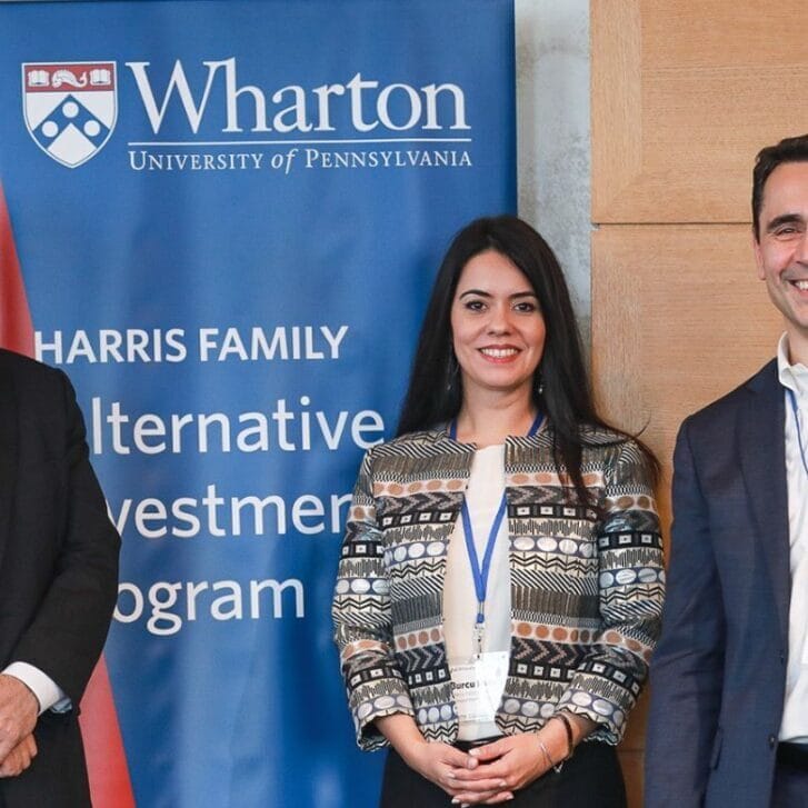 Two men and a woman in business attire stand in front of a sign that says Harris Family Alternative Investments Program and has a Wharton logo at the top of it.