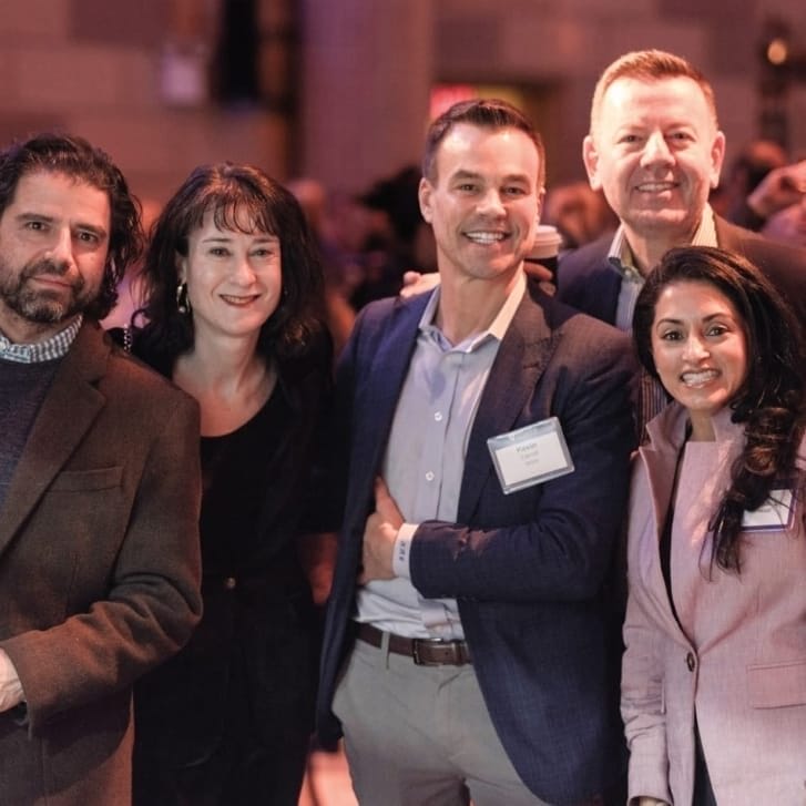 Six people in business clothing gather for a photo in a crowded event space.