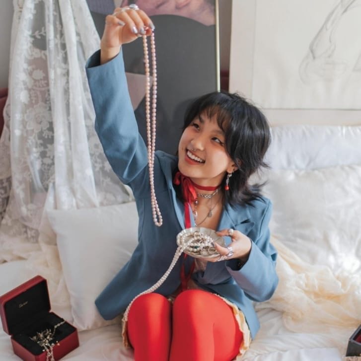 A woman in a blue suit jacket, red leggings, and silver shoes sits on a bed with books and jewelry around her and holds up a pearl necklace.