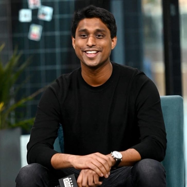 Portrait of Ankur Jain wearing a black shirt and black jeans seated on a chair and holding a microphone.