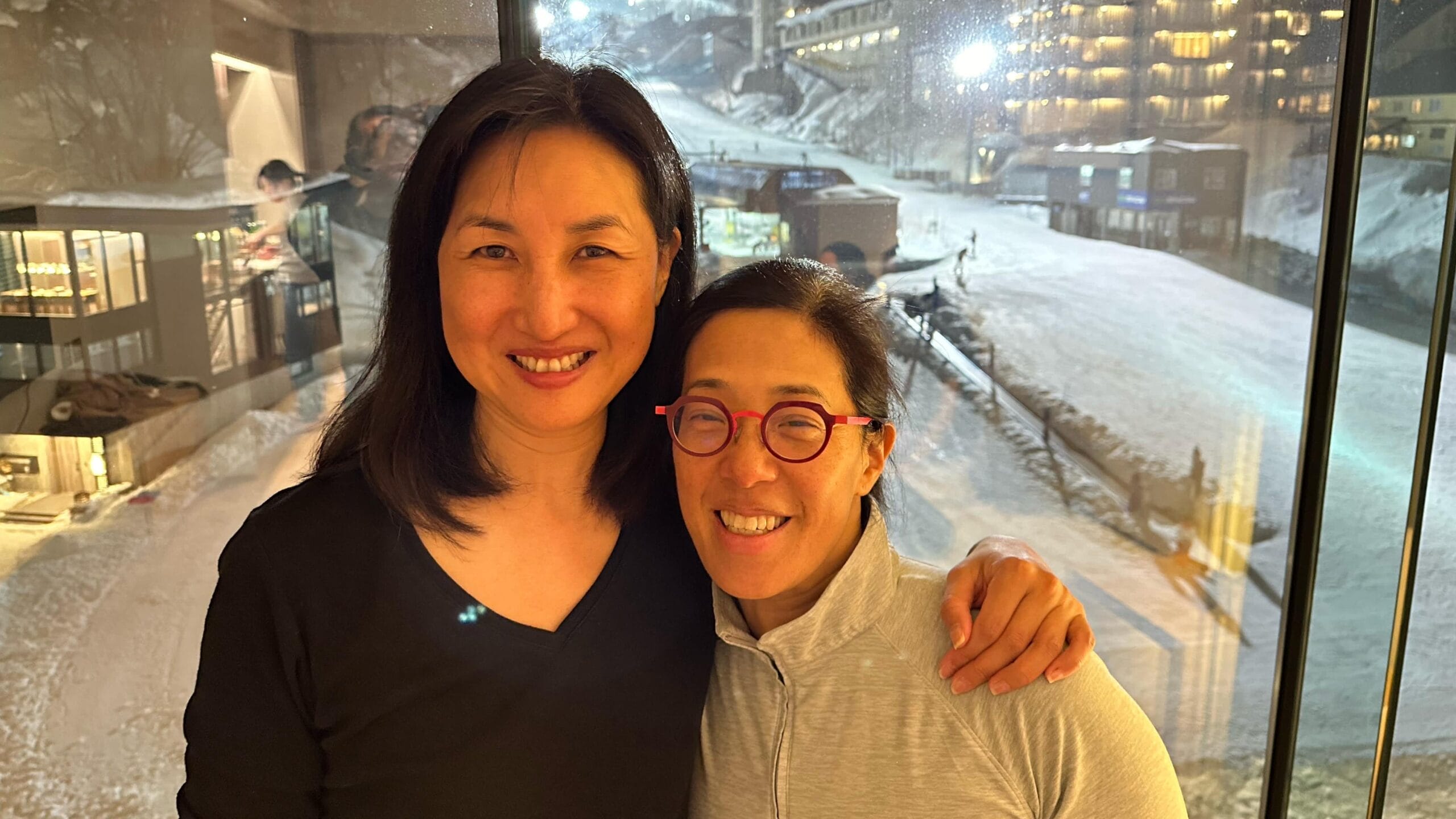 Two women pose together for a photo against a window with snow on the ground outside.