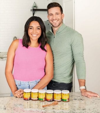 A woman in a pink shirt and a man in a light green shirt stand together behind a marble countertop that has six jars of coconut spread on it.