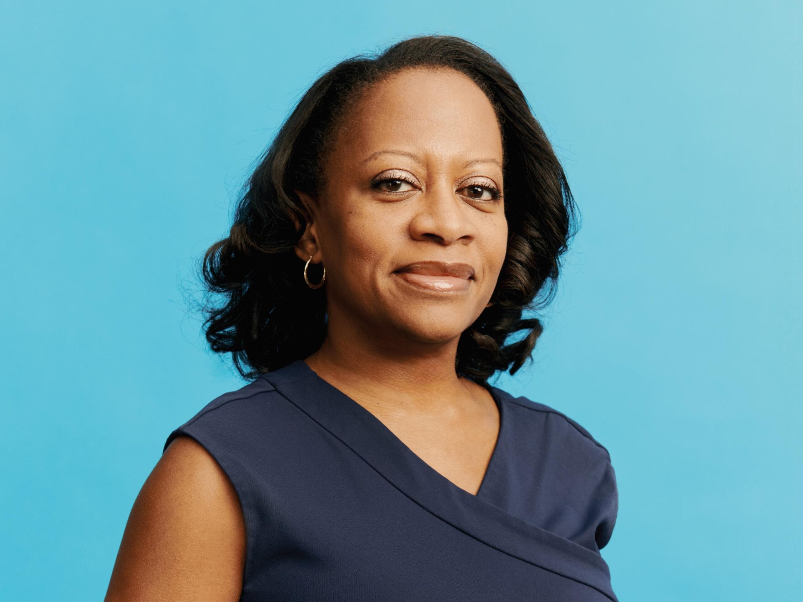 Woman in a dark blue dress and with curled black hair poses for a photo against a bright blue backdrop.