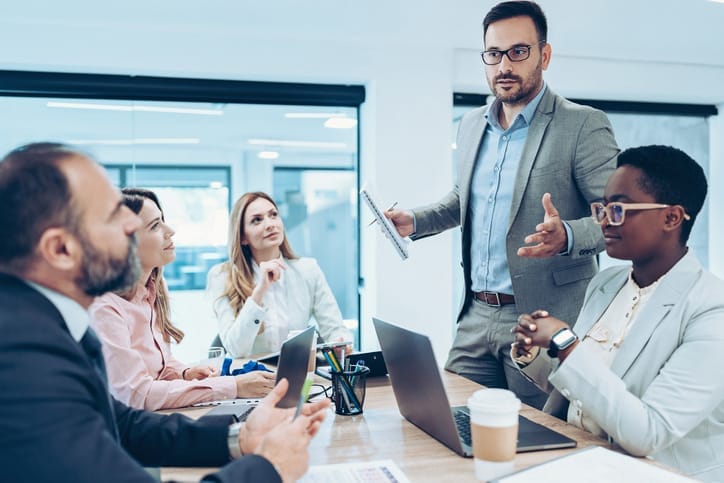 Man standing in a board room, gesturing widely in front of colleagues