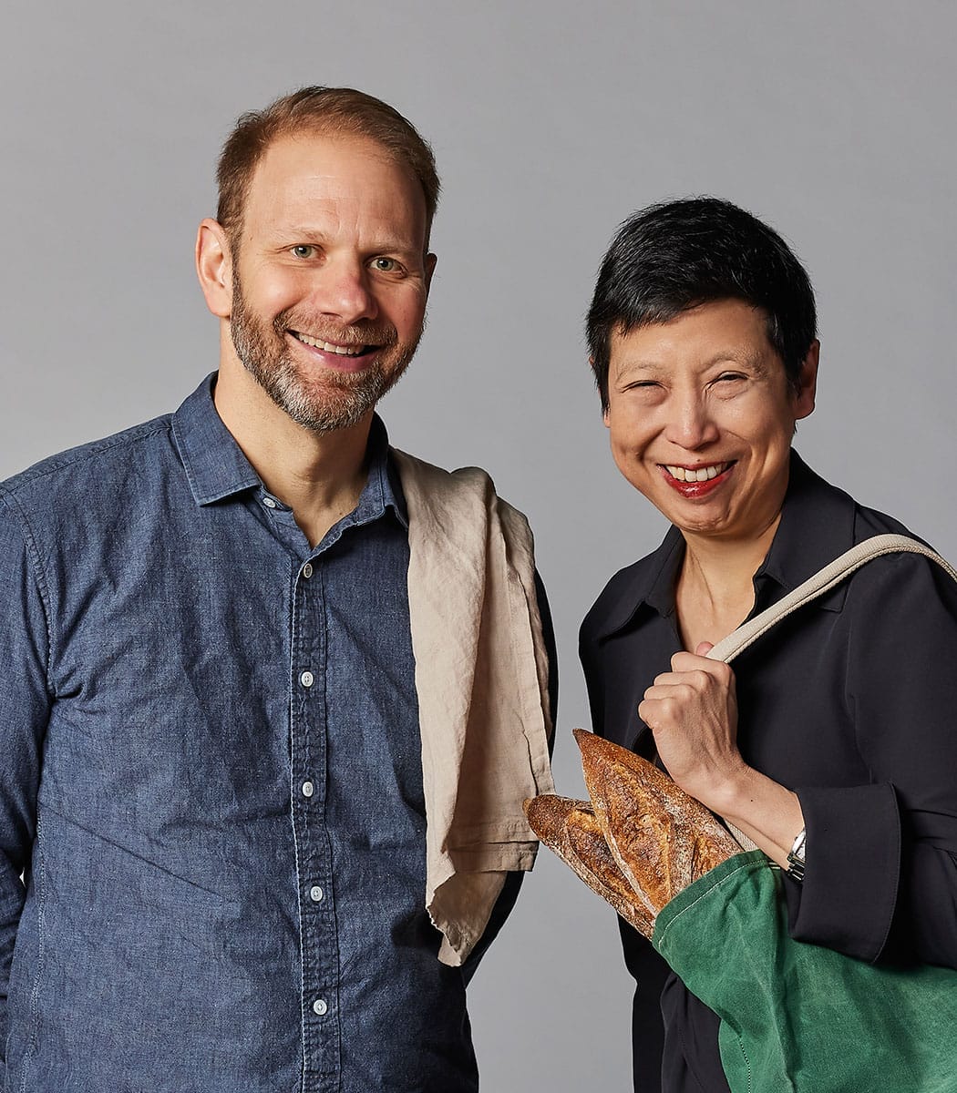 A man in a denim button-down shirt and a woman in a black button-down shirt pose together, a tan towel draped over one of the man's shoulders and a green bag with bread in it draped over one of the woman's shoulders.