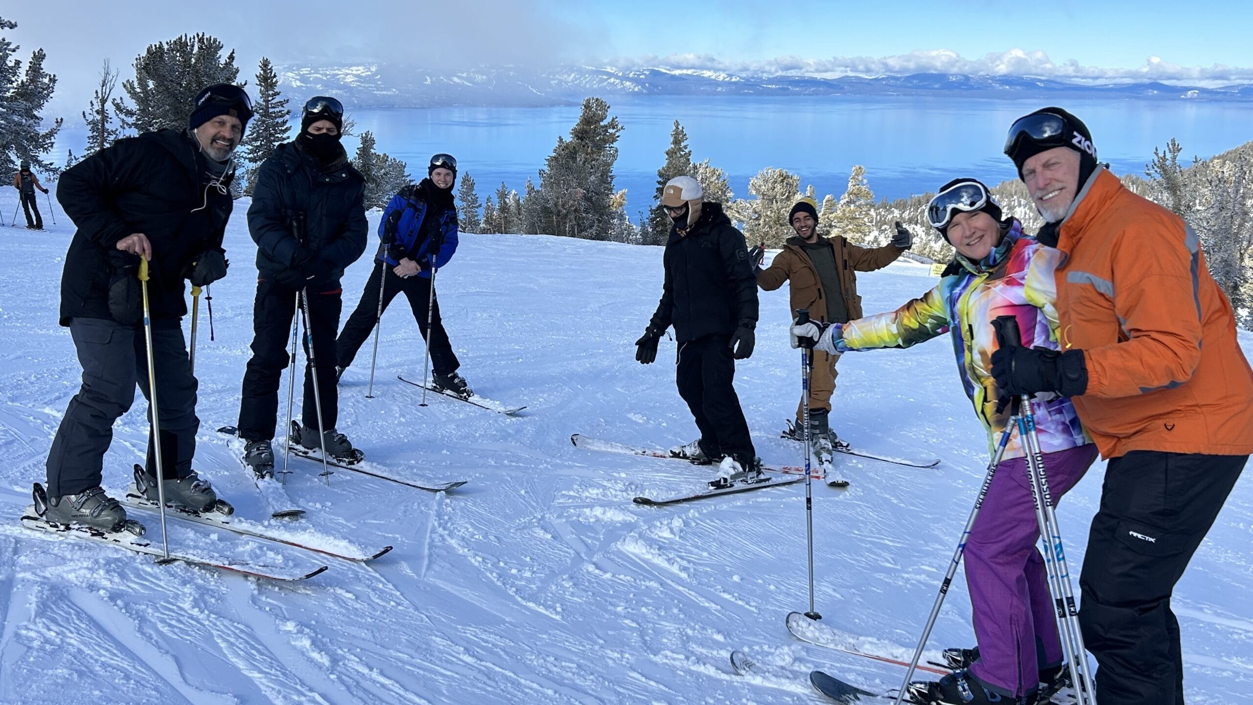 Seven people pose for a photo atop a mountain, all on skis.