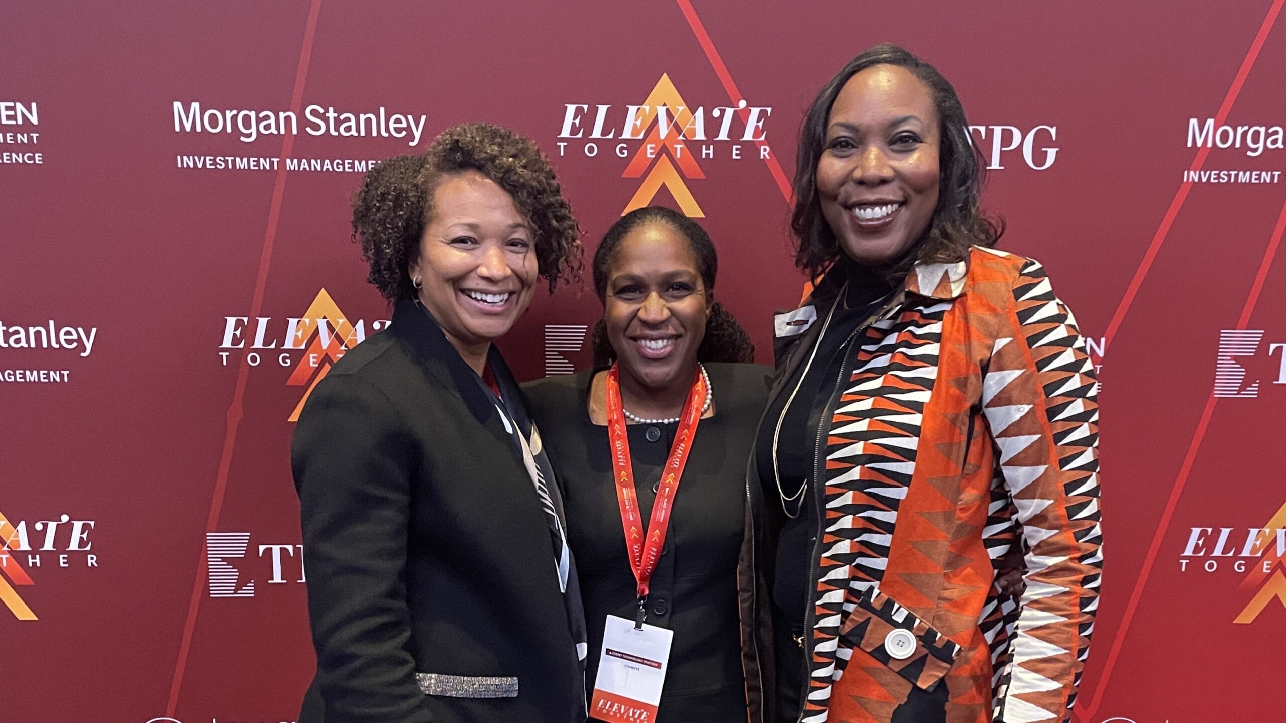 Three women in business attire pose together for a photo in front of a red backdrop.