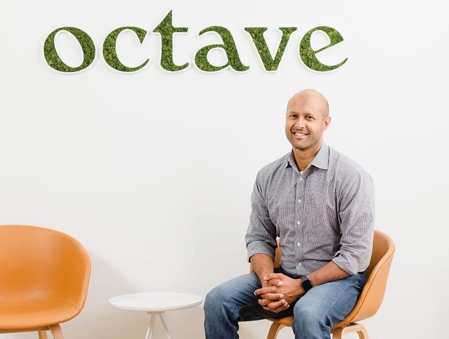 Sandeep Acharya in jeans and a button-down shirt seated in front of a wall with an Octave logo on it.