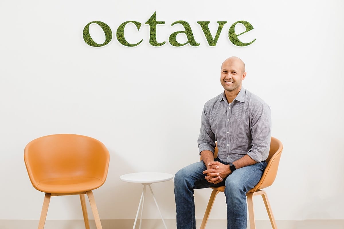 Sandeep Acharya in jeans and a button-down shirt seated in front of a wall with an Octave logo on it.