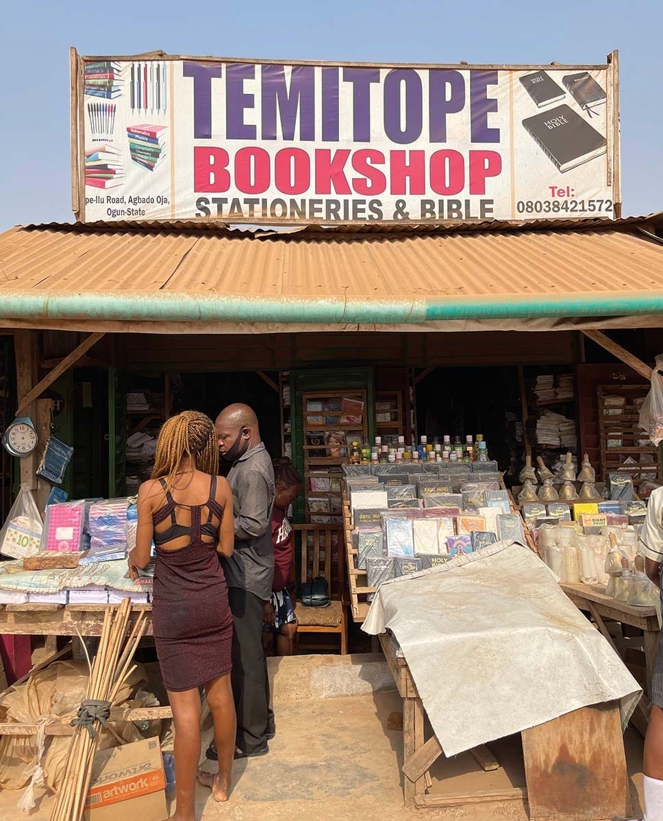 An open-air bookshop with a sign on the roof with the name Temitope Bookshop and advertisements for stationary and bibles.