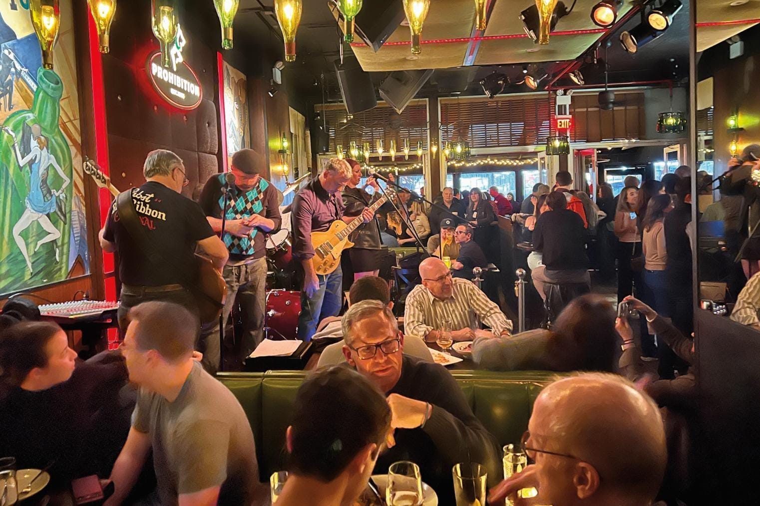 Two guitarists and other band members get ready on stage in a bar as people eat and drink around them.