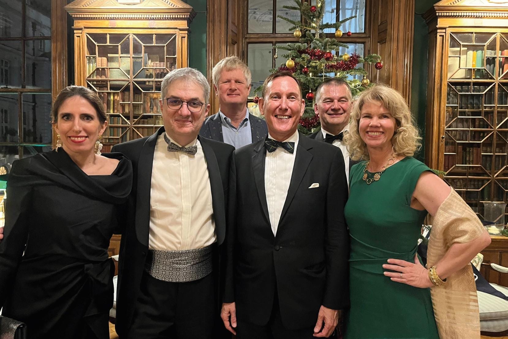 Six people in formal attire stand in front of two ornate bookcases and a Christmas tree.