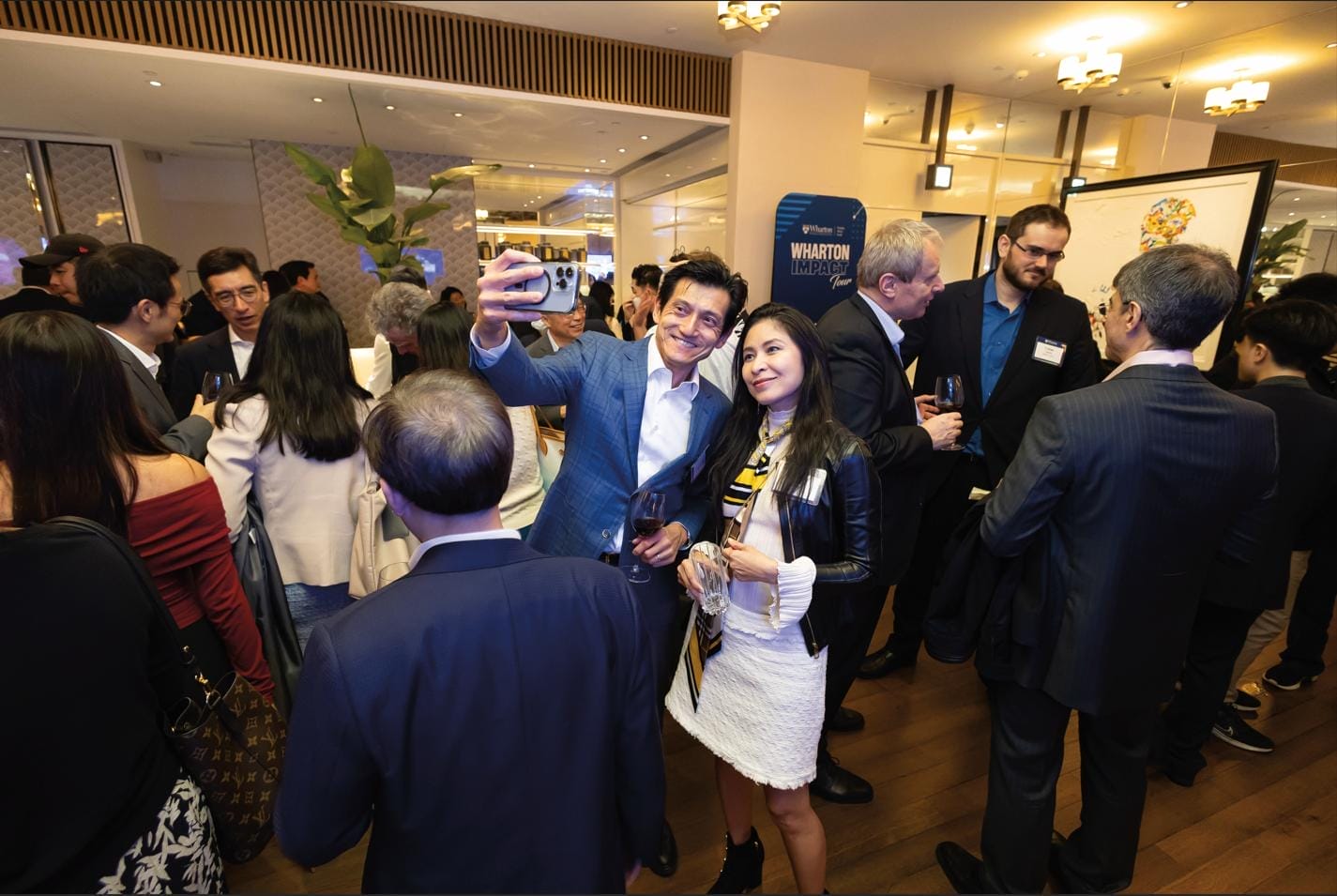 Two people in business attire take a selfie in a room filled with mingling people.