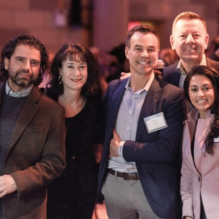 Six people in business clothing gather for a photo in a crowded event space.