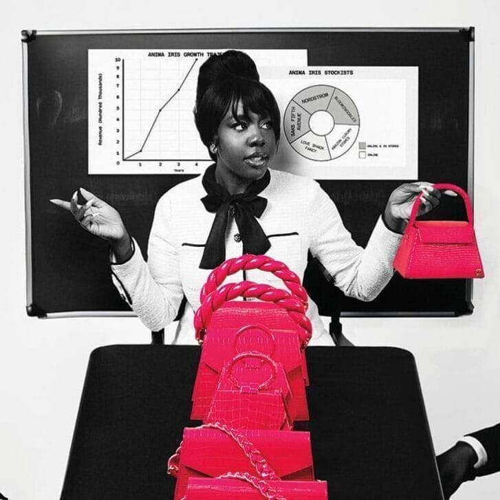 Wilglory Tanjong in a white jacket and with a black bow around her neck stands in front of a whiteboard holding a bright pink bag. Other bright pink bags are arranged on a table in front of her.