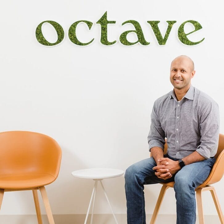 Sandeep Acharya in jeans and a button-down shirt seated in front of a wall with an Octave logo on it.